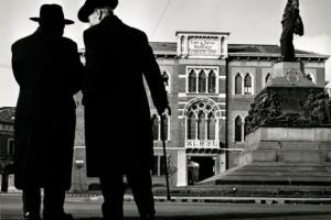 Two men in winter coats and hats in the style of the early 20th century speak to one another closely before a large courtyard with a statue at its center. 