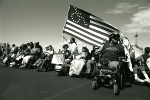 A group of protesters, many using wheelchairs, with an American flag with an icon of a person in a wheelchair instead of the usual stars.