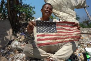 An Indonesian man holds a tattered American flag in a trash heap. 