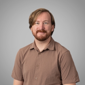 A white man with strawberry blonde hair and a short beard smiles at the camera against a grey backdrop. He wears a brown collared shirt.