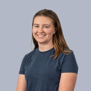 A woman with dirty blonde hair and blue eyes smiles to the camera against a gray backdrop. She wears a navy blue t-shirt.