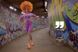 A colourful drag queen, wearing an orange foam wig, a purple leotard and high heels, marches through a Belfast tunnel whose walls have been spray-painted.