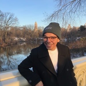 A person with a beanie stands in front of a body of water during the winter. They wear a white shirt and a black pea coat.