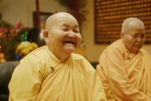 Two Buddhist nuns in a temple, with shaved heads, and goldish robes. The Buddhist Nun on the left is smiling big.