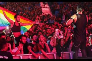 A singer in a leather outfit stands before a crowd of fans, one waving a Pride Flag over their shoulders.