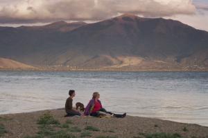 Two women with short hair, and a dog, sit in front of a lake and mountains