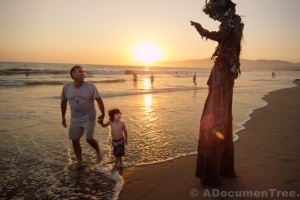 A man on stilts dressed as a tree waves to little boy and his father on the beach at sunset