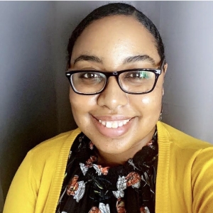 A woman with glasses, black hair, black eyebrows and brown skin smiles at the camera in a selfie style photo. She wears a button up shirt with a pattern and a mustard-yellow cardigan.