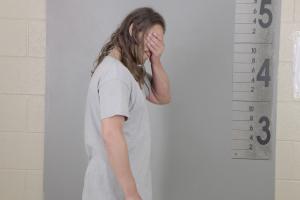 A Caucasian man with long hair covers his face with his hand while standing in front of a mugshot background 