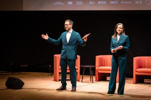 Two white people in navy blue suits stand on stage.