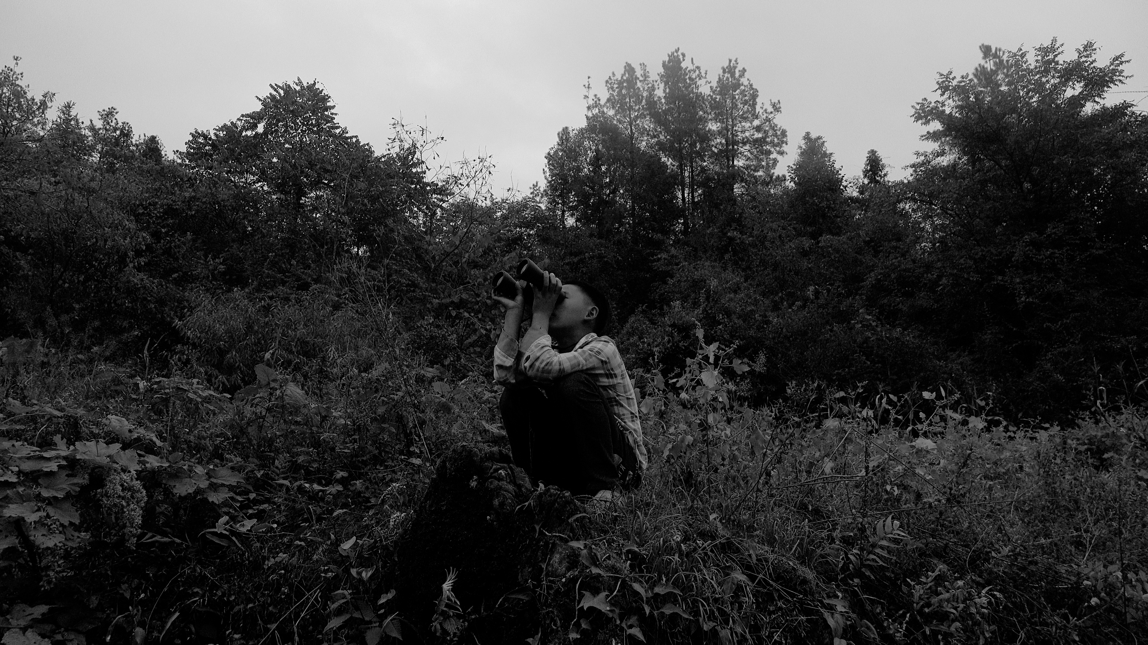 A person squating in a field, looking through binoculars.