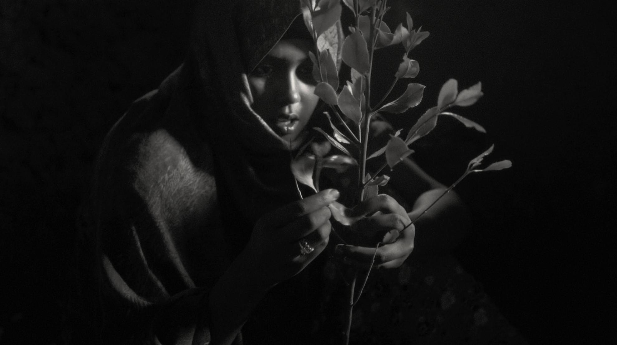 Black and white image of Habu Abdella, a young Ethiopian woman holding a stem of khat leaves. From Jessica Beshir's 'Faya Dayi.' Courtesy of Feyatey LLC