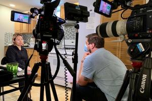 A light-skinned man wearing mask is sitting behind two large cinecameras on tripod, filming a Latino woman in the kitchen.