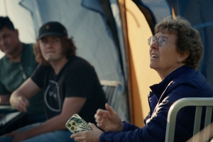 During golden hour, an older bespectacled astronomer looks up anxiously to the sky. Her younger colleagues, at a distance in a tent behind her, look to her. All are concerned.  