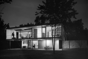 A black and white photo from the 1960's of a mid-century modern house at night.  It's an "upside down" house where the second story is larger than the first story so the house looks like two elegant rectangles stacked atop each other. The house is lit from the inside and a tall, daper man in a tuxedo stands on the upstairs terrace. A woman sits inside on the living room couch, and down below two young children can be seen.