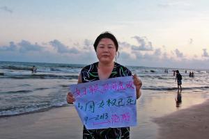 Still image from Nanfu Wang’s "Hooligan Sparrow." Ye Haiyan, a fair complexioned adult with black hair, stands at the beach wearing a black dress with blue, green, and white designs. She is holding a sign with simplified Chinese characters.