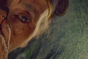 Still image from 'The Mother of All Lies,' showing a closeup shot of half of an old woman's face. She is holding a figurine covering her mouth.