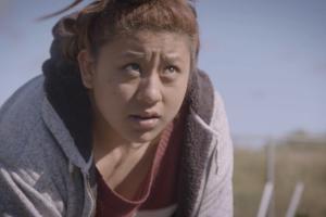 A Latina teenager leans over in a field, tilting her head upwards and looking away from her harvesting work.
