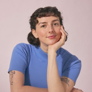 A woman with short black hair sits with one hand on her face and one resting on the table, smiling. It has the vibe of an old Hollywood portrait. The background is mauve.