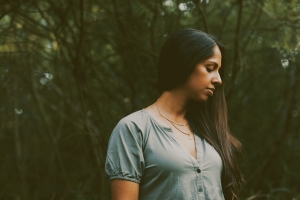 A dark-skinned woman with long black hair wearing a sage green dress looks downward as she is surrounded by trees.