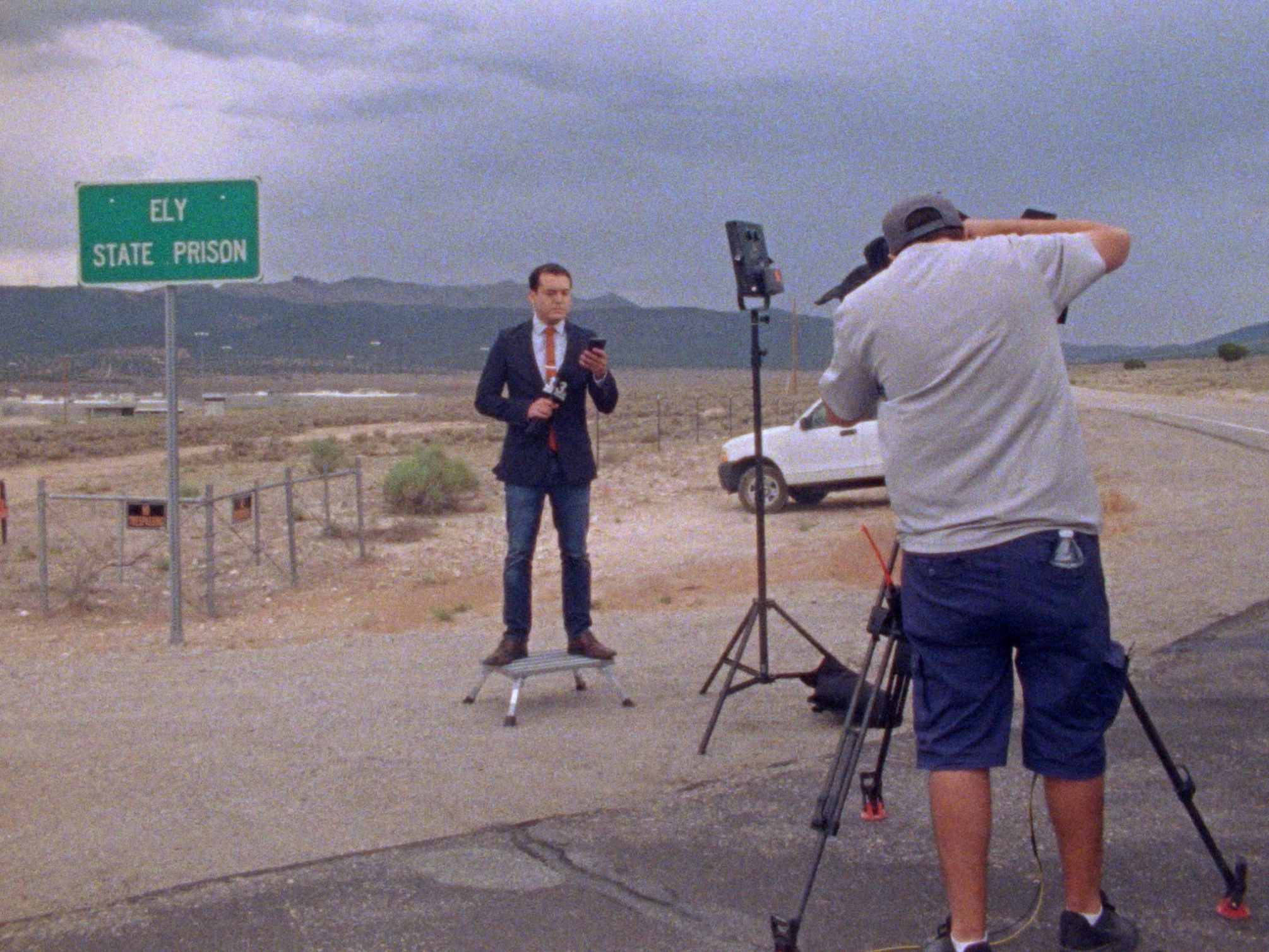 A news anchor stands in the middle of the desert, in front of a sign that says, “ELY STATE PRISON”. A production light shines on them, and a camera person with a tripod films them. 