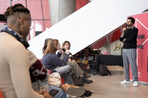 In a bright room with concrete pillars and floors, a man in a sweater answers a question from the audience.