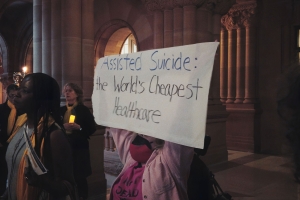A woman in a pink jacket holds up a sign that reads: "Assisted Suicide: The World's Cheapest Healthcare."