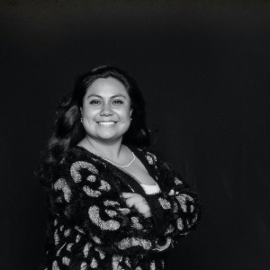 Black and white photo of a woman with long, wavy black hair smiling brightly at the camera. She crosses her arms confidently. She wears a patterned long and long-sleeved dress.