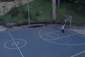 An African American man wearing a gray hoodie stands on an empty basketball court, reaching up to touch the basketball net