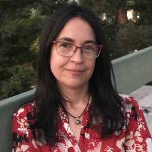 A woman with black hair and red-framed glasses smiles at the camera in an outdoor area. She wears a red blouse.