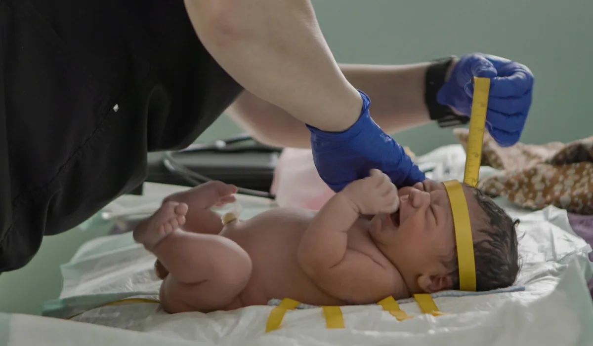 Film still from 'Arrest the Midwife'. A newborn baby being measured by a person with blue gloves and black scrubs.