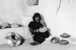 Nampeyo sits alone in a room surrounded by her masterpieces. Seemingly unaware of the photographer, she gazes introspectively at a ceramic seed jar in her lap.
