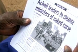 Image of two hands holding a folded newspaper with the headline "Acholi leaders to cleanse ministers of naked curse"