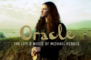 A middle aged man with long hair and a guitar stands on the coastal bluffs of California.