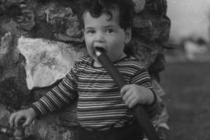 A white male toddler stands in front of a stone wall, chewing on a garden hose.