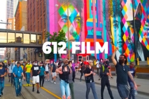 Thousands of protestors dressed in summer clothes march in front of a colorful mural 