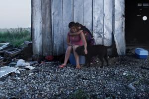 two female siblings sit on a plastic box and pet their black dog in front of a rural dilapidated barn.