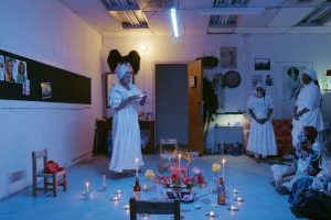 Still of a group of people in white dresses, conducting a ritual in a darkened studio.