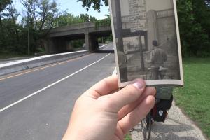 A hand holds an old photograph of a man with a bicycle at South Amboy Bridge. The photo lines up perfectly with South Amboy Bridge in the background. 