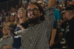 Film still from 'Your Friend, Memphis,' depicting a smiling young white man in a checkered shirt, raising his left arm, in a crowded stadium.