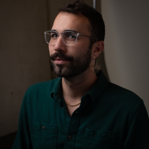 Headshot of Zaferhan Yumru, a queer male presenting person with short black hair, wearing transparent eyeglasses, a gold earring in the shape of an eye, a gold chain necklace and green shirt.