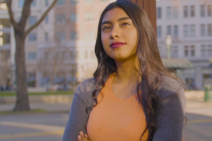 A young Latina woman stands with arms crossed in front of distant buildings