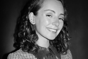 Headshot of Berry Hahn. A black and white image of a smiling mixed-race woman with mid-length curly brown hair, wearing a jacket with a houndstooth pattern.
