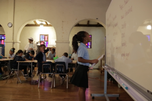 a grade school African American girl in a school uniform looks up at a white board with a paper in her hand in front of the rest of her young classmates.