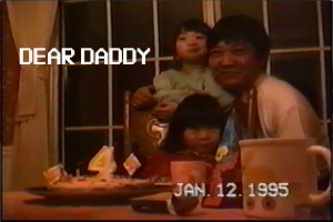 Screenshot of a VHS tape recording of a father and his two young children sitting at a table, in front of a birthday cake with a number four candle on it. The date on the tape reads Jan 12 1995.