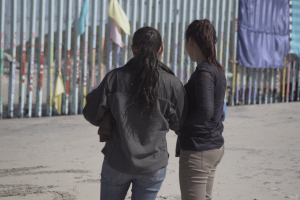 Photographed from behind, two people with long black hair stand in front of a tall, grey border wall.