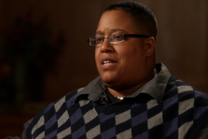 A middle-aged black individual in a patterned sweater sits in front of a muted brown backdrop