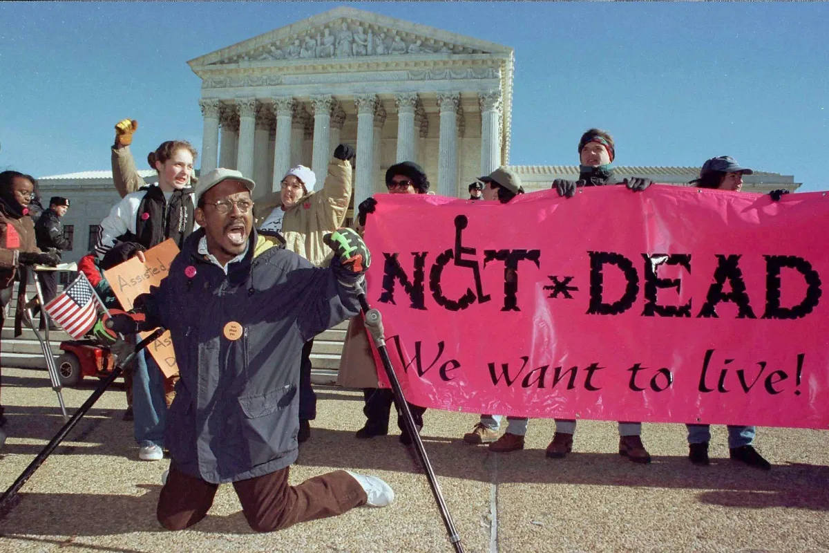 People protesting. In front is a Black disabled man. Behind him is a hot pink banner with black text that reads 'Not Dead. We want to live!'