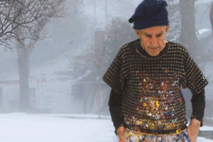 an older White Man with a paint-splattered sweater and knit cap stands in front of a snow-covered house.