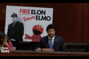 Pixelated screen shot of a U.S. congressman in front of a "Fire Elon Save Elmo" sign. The PBS News logo is in the bottom left corner.